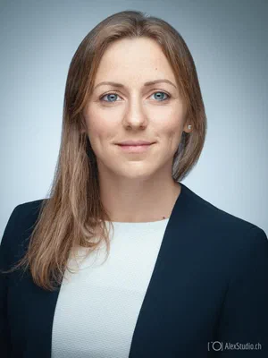 Polished headshot of a poised professional in a dark blazer and white top, softly lit against a clean pale blue studio background.