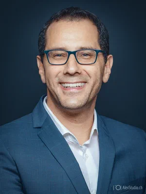 Warm LinkedIn profile photo of a smiling professional in blue glasses and a tailored blue suit, photographed against a dramatic dark backdrop.