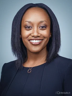 Bright professional profile featuring a welcoming smile, understated jewelry, and a dark blazer, captured with balanced lighting on a pale blue studio backdrop.