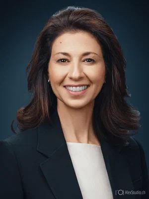 Welcoming corporate photo with a broad smile, dark blazer, and cream top, photographed with luminous skin tones against a rich blue backdrop.