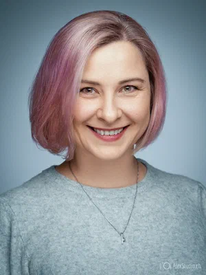 Friendly photo for a resume or CV featuring a cheerful professional with a pink bob, grey knit top, and delicate necklace on pale blue.