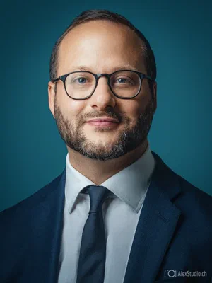 Distinguished executive portrait featuring glasses, a neatly trimmed beard, navy suit, white shirt, and tie, with directional light across a teal background.