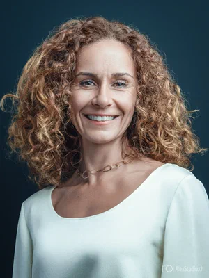 Expressive corporate portrait with a bright smile, voluminous curls, white scoop-neck top, and gold necklace, created by Alex, Photographer based in Geneva, Switzerland.