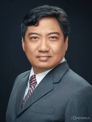 Formal headshot of a composed professional in a pinstriped grey suit and checked tie, precisely lit against a clean dark studio background.