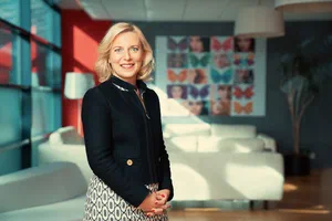 A bright editorial portrait of a smiling woman in a black jacket and patterned skirt, photographed by Alex in a contemporary Geneva office.