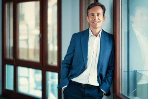 Relaxed business profile image of a smiling man in a blue suit, leaning beside tall windows in soft natural light.