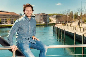 Casual environmental portrait of a smiling man in denim, seated on a waterfront railing with city architecture and blue sky beyond.