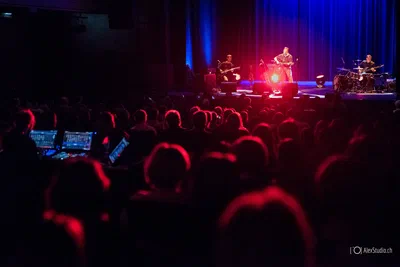 A packed audience faces a live band under vivid red and blue stage lighting, captured in atmospheric event photography from behind the sound desk.