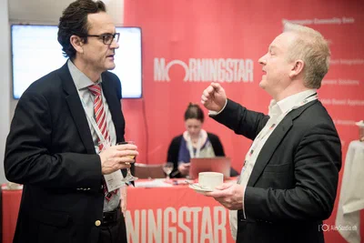 Two suited attendees share an animated conversation beside an exhibition stand, a candid networking moment photographed with crisp, polished corporate event style.