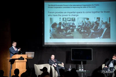 A presenter speaks from a lectern beneath a projected forum slide while seated panelists listen, creating a strong conference photography composition.