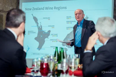 A lecturer presents a map to seated guests, with glassware and bottles in the foreground adding depth to this professional event photograph.