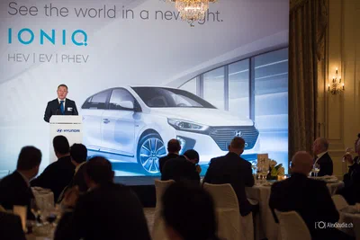 A suited speaker addresses a seated dinner audience before a large automotive presentation screen in elegant, warmly lit conference surroundings.