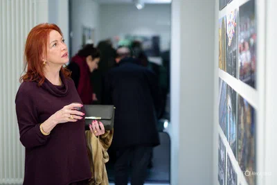 A woman pauses with a drink to study a wall display while other reception guests mingle softly out of focus behind her.