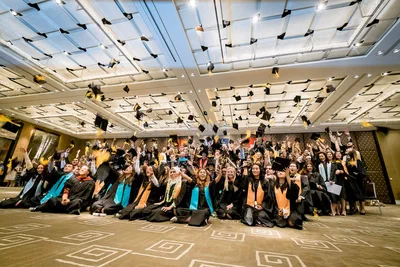 A large group celebrates together in a hotel ballroom, raising hands beneath geometric ceiling panels for an energetic corporate event portrait.