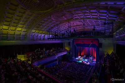 A sweeping balcony view reveals a full theatre and performers on a red-lit stage beneath dramatic purple architectural lighting.