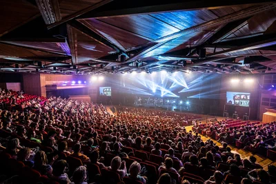 Hundreds of attendees fill a vast auditorium as brilliant stage beams cross overhead, creating an expansive, high-energy conference photography scene.