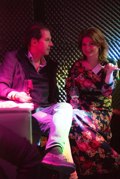 Two women converse closely while seated under magenta and blue lighting, an intimate candid from a relaxed evening event setting.
