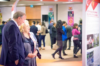 Two visitors read an informational display as groups network throughout a bright exhibition hall, documented with clean corporate event photography.