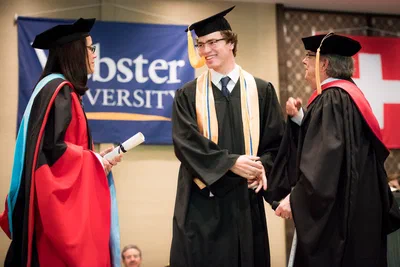 A smiling graduate in cap and gown shakes hands onstage between academic officials, captured during a formal ceremony by Alex in Geneva.