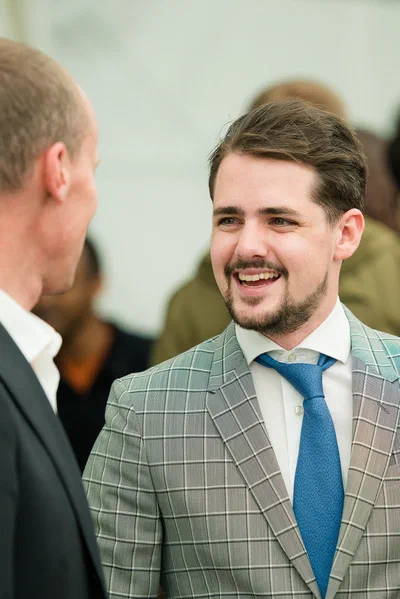 A man in a checked suit smiles mid-conversation, isolated against a softly blurred crowd in refined candid corporate photography.