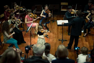 A young violin soloist performs beside a conductor and seated string ensemble, captured in warm stage light with the audience framing this event photograph.