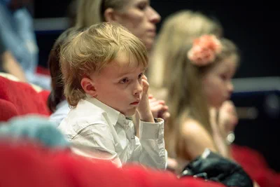 A young audience member watches intently from a red theater seat, isolated against softly blurred guests in this candid event photography moment.