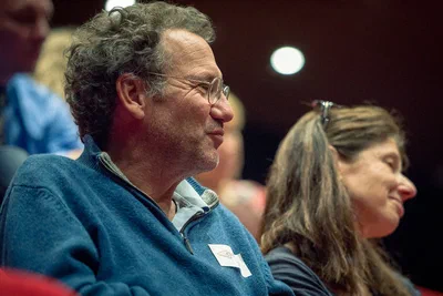 A bespectacled attendee smiles while listening in a theater, with shallow focus lending this conference photograph a warm, attentive atmosphere.