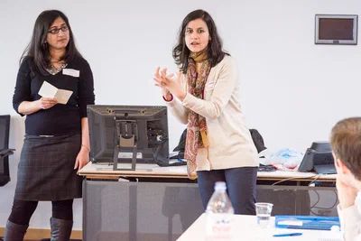 Two women lead a bright workshop session beside a desktop monitor, one gesturing expressively as seated participants follow the corporate presentation.