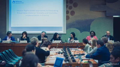 Panelists and delegates gather around a formal conference table beneath a projected human-rights presentation, documented by Alex, a photographer in Geneva, Switzerland.