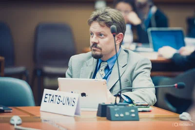 A focused delegate sits behind an ETATS-UNIS placard with a tablet, microphones, and translation equipment in a composed conference photography portrait.