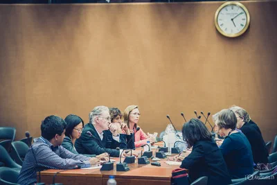 Delegates confer across a microphone-lined table beneath a wall clock, creating a balanced documentary view of a formal international meeting.