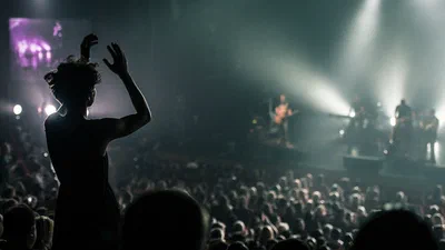 A silhouetted audience member dances above a packed concert crowd while brilliant stage beams cut through haze toward distant performers.