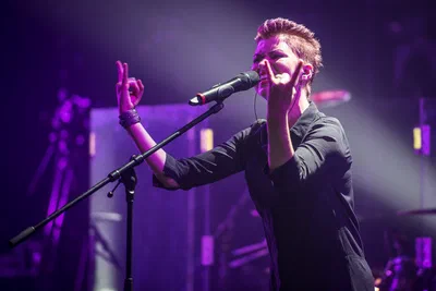 Purple stage light surrounds a singer performing passionately at the microphone, hands raised in an expressive live-event photography close-up.