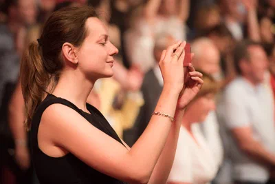 A woman records the occasion on her phone amid a softly blurred crowd, captured with warm light and candid event-photography intimacy.