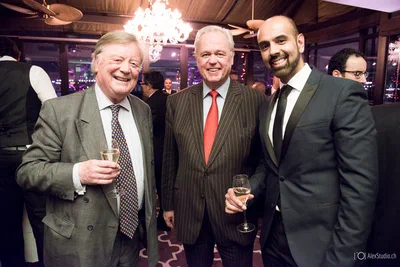 Three suited guests smile together over champagne at an elegant evening reception, framed beneath chandeliers in polished corporate event photography.