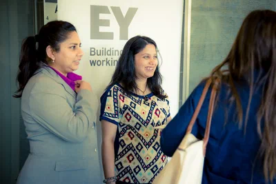 Three attendees share a lively conversation beside prominent EY signage, conveying the approachable energy of a professionally photographed networking event.