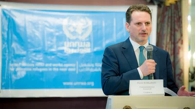 A suited speaker addresses guests with a handheld microphone before a blue UNRWA backdrop in crisp, focused conference photography.