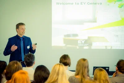 A presenter gestures beside a projected “Welcome to EY Geneva” slide while a full audience listens, photographed in a bright corporate setting.