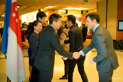 Two suited guests exchange a firm handshake beside Singapore flags as others mingle behind them at a warmly lit corporate reception.