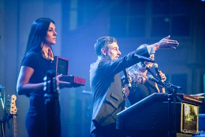A host gestures toward the audience from a blue-lit podium while two women stand nearby with microphones and an open presentation box.