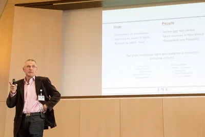 A conference speaker presents a projected comparison of dogs and people, holding a clicker against the clean lines of a modern lecture room.