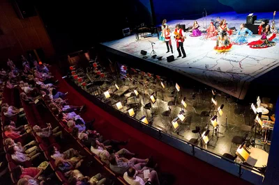 Colorfully dressed performers take the stage above an empty orchestra pit as theater guests watch, photographed from a dramatic elevated viewpoint.
