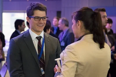 Two conference attendees share a friendly conversation amid a softly blurred networking crowd, with cool ambient lighting and an approachable corporate atmosphere.