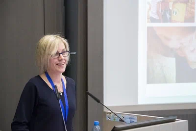 A smiling speaker addresses the room from a lectern beside a projected slide, creating a bright, engaging moment of professional conference photography.