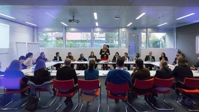Attendees gather around a wide U-shaped table while a standing presenter leads the discussion in a modern, daylight-filled corporate meeting room.