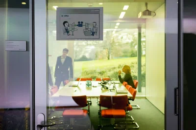 Seen through a glass door, two participants meet around a long conference table, framed by orange chairs, clean architecture, and garden views.