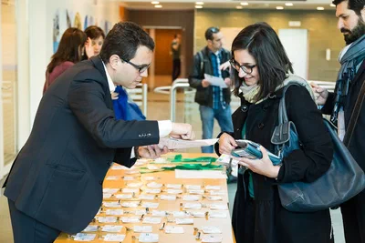 A staff member assists a smiling attendee at a busy registration table covered with badges, capturing the welcoming energy of a conference arrival.