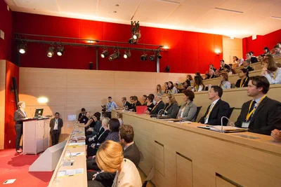 A lecturer speaks from the front of a tiered auditorium as attendees follow attentively, framed by warm wood, red walls, and stage lights.