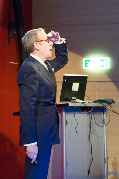 A suited presenter gestures animatedly beside a lectern and monitor, delivering a lively conference talk in a clean, tightly framed vertical composition.