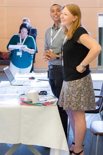 Two badge-wearing participants laugh together beside a resource table while another attendee takes notes, conveying an informal and collaborative workshop atmosphere.